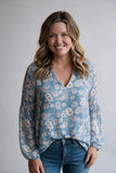 A woman with wavy blonde hair smiling, wearing a light blue long-sleeve blouse with a white and beige floral pattern, paired with blue jeans, against a neutral gray background.