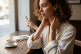 A woman with curly brown hair sits at a wooden table in a cafe, wearing a cream-colored linen button-up shirt and gold geometric earrings. She is looking thoughtfully to the side, with a coffee cup on the table in the foreground.