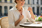 A woman in a white blouse sits at an outdoor cafe table holding a white cup, wearing a chunky amber and cream beaded necklace. The background shows a softly blurred street scene with buildings and flowers.