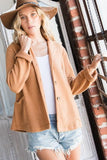 A woman wearing a camel-colored button-up cardigan, a brown wide-brim hat, a white tank top, and denim shorts, posing in front of a rustic brick and window background.