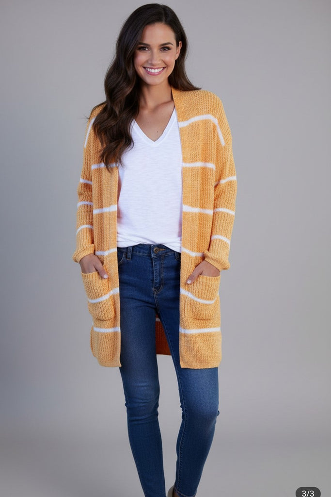 A woman modeling a long, open-front ribbed cardigan with horizontal white stripes on a mustard yellow base, worn over a white V-neck top and blue jeans against a solid gray studio background.