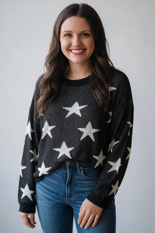 A woman wearing a dark charcoal gray knit sweater with a white star pattern, paired with blue jeans, standing against a light gray background.